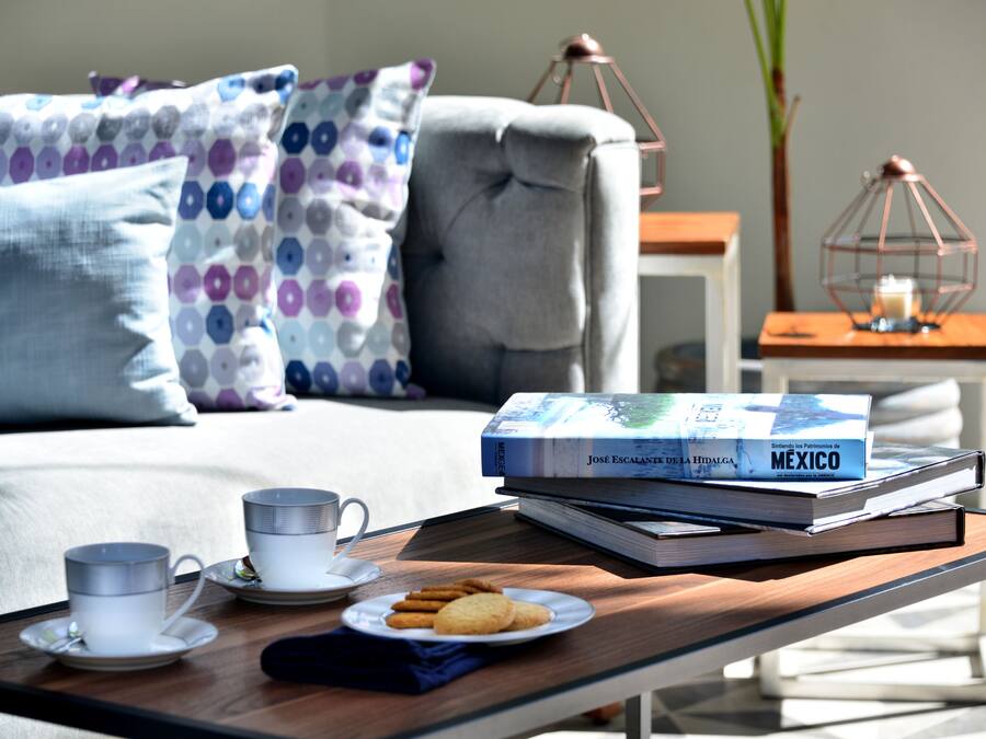 Una sala acogedora con un sofá, almohadas, una mesita de centro con un libro y galletas, y una mesita auxiliar con una vela.