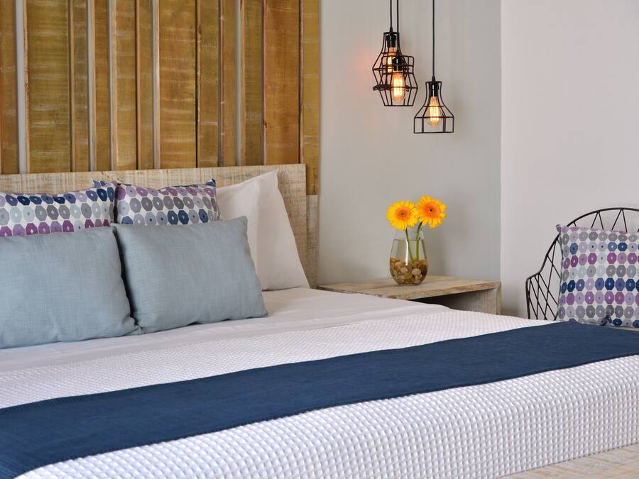 Una cama bien hecha con edredón azul y blanco, dos almohadas grises y una almohada estampada. Hay una mesita con un jarrón de flores amarillas y un cuenco de nueces.