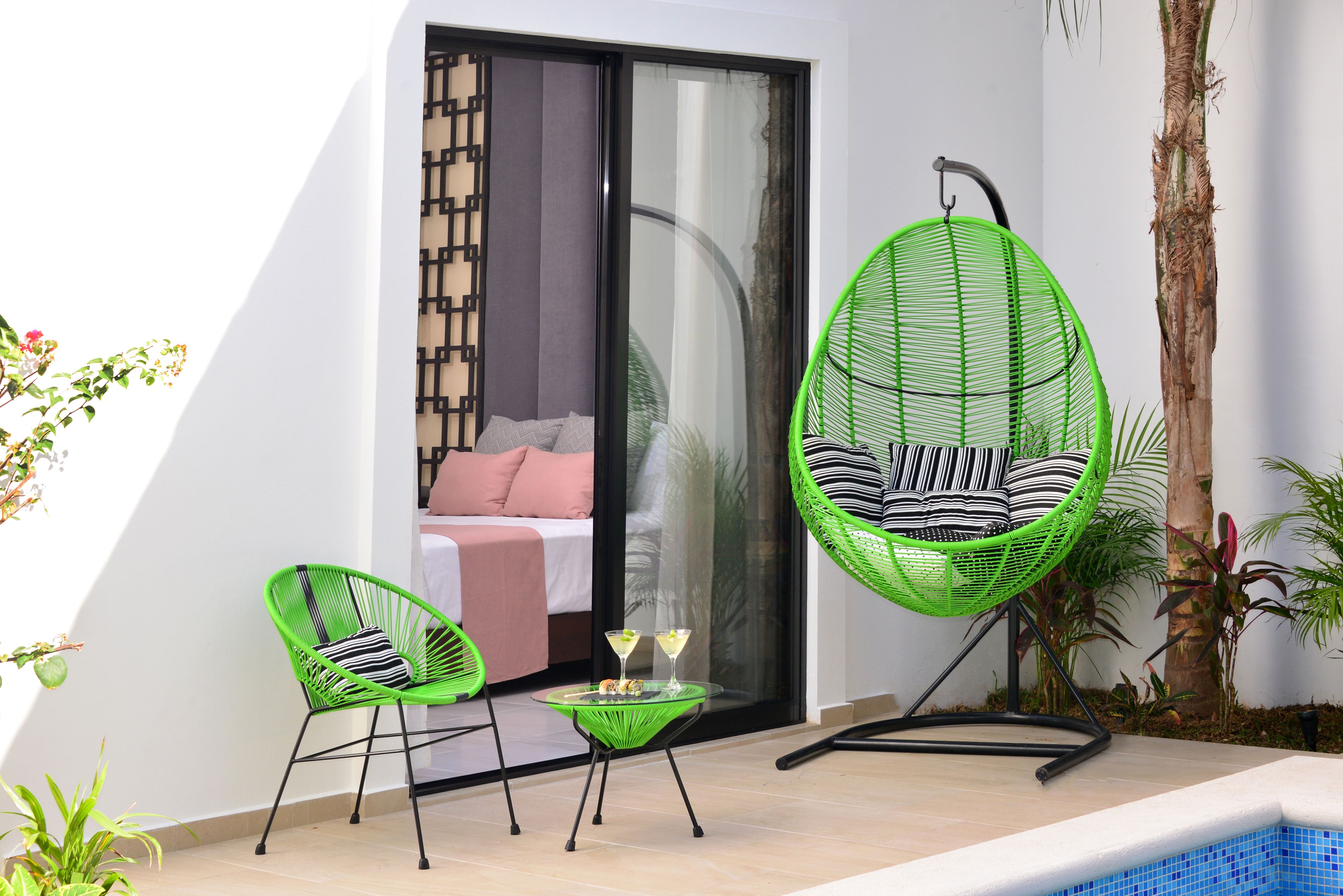 A modern outdoor seating area with two green chairs and a glass table, a sliding glass door leading to a bedroom, and a small pool with blue tiles.
