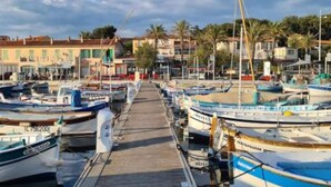 A marina with boats docked along a wooden pier, leading to a town with buildings and palm trees. - Holiday Home in Villecroze with Pool & Garden (Villecroze)