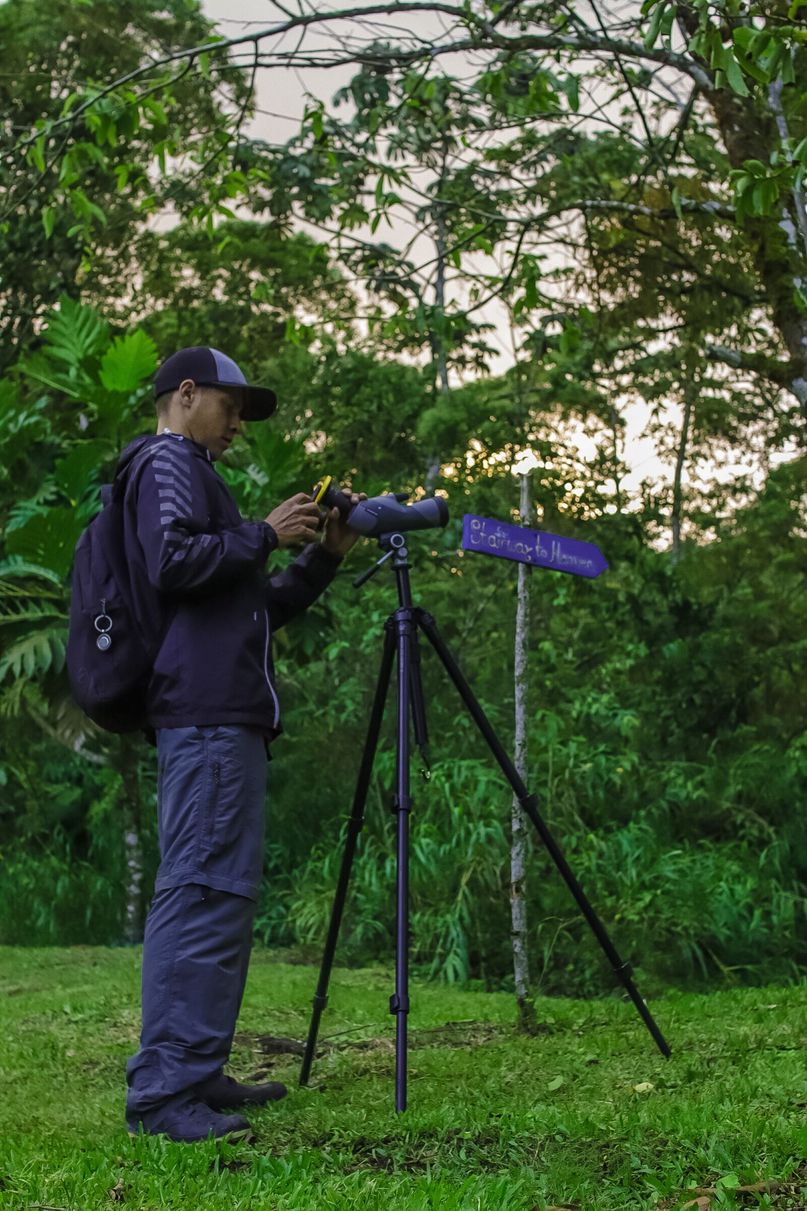 Une personne ajuste un appareil photo sur un trépied, dans un environnement naturel, avec de la végétation et un panneau indicateur en arrière-plan.