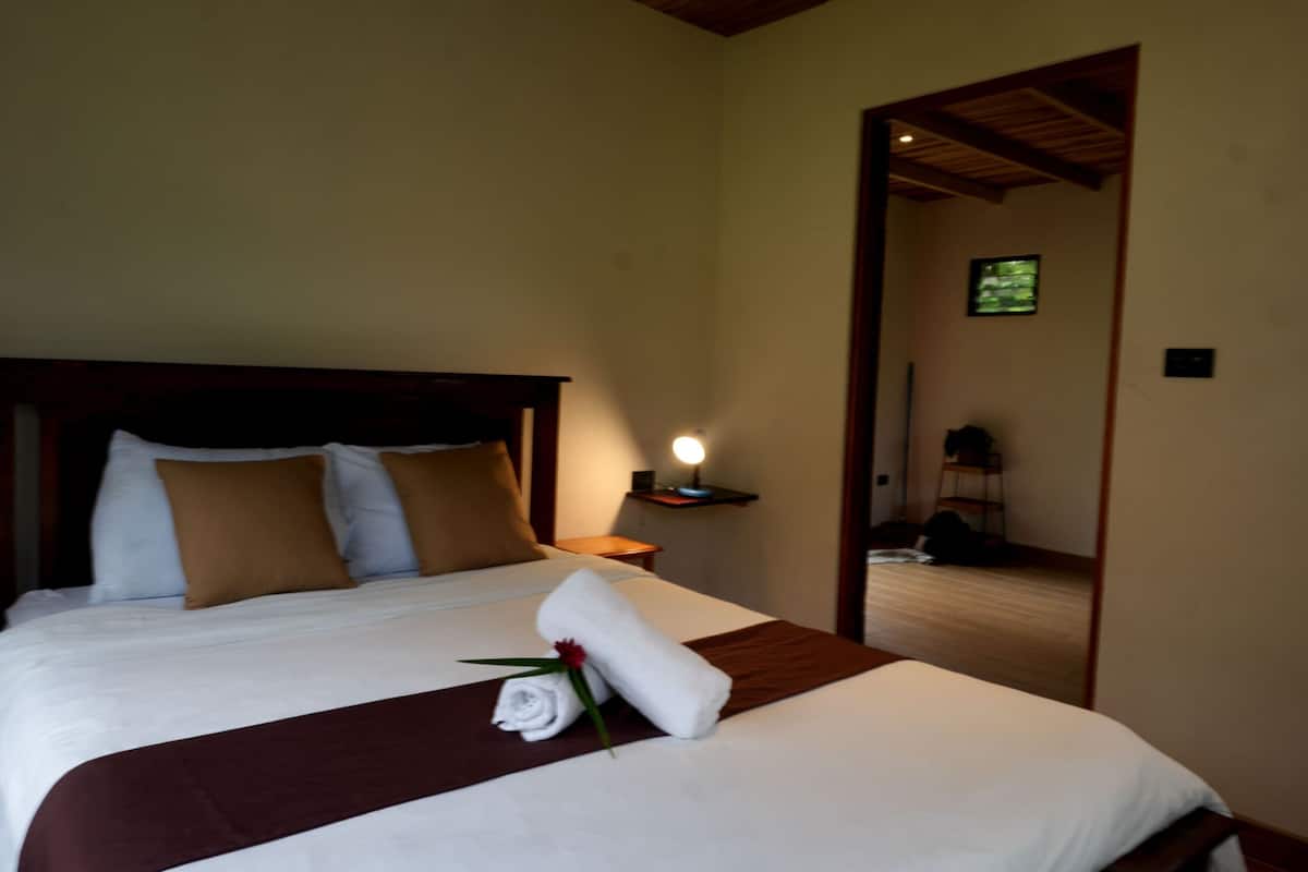 A bedroom with a wooden ceiling, a large bed with white linens, a brown throw blanket, and a bedside lamp.