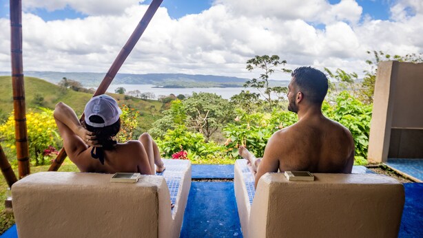 Two lounge chairs facing a pool with a view of a lake and hills.