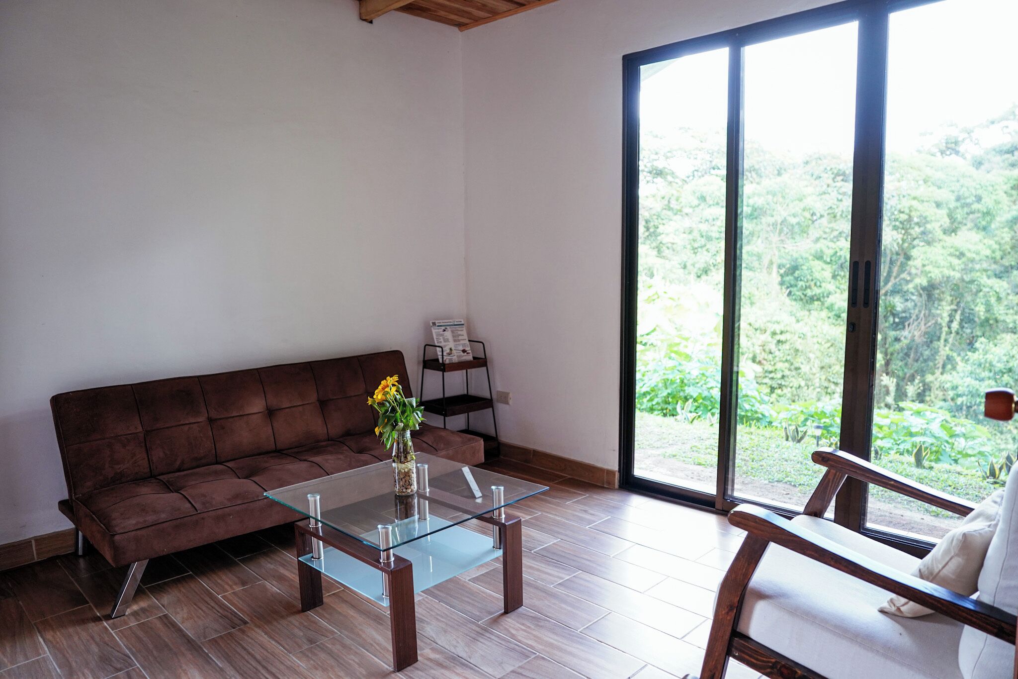 A modern living room with a brown leather sofa, a glass coffee table, and a view of a lush green landscape through large sliding glass doors.