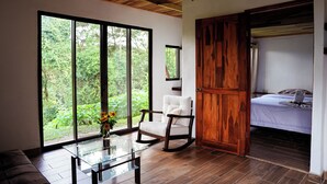 A room with a large sliding glass door leading to a garden, a wooden rocking chair, a bed with a white comforter, and a glass coffee table with a vase of flowers.
