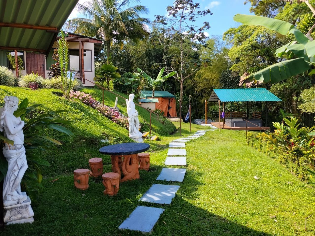 Um jardim com uma estátua, uma mesa com bancos, um caminho e um edifício com telhado verde.