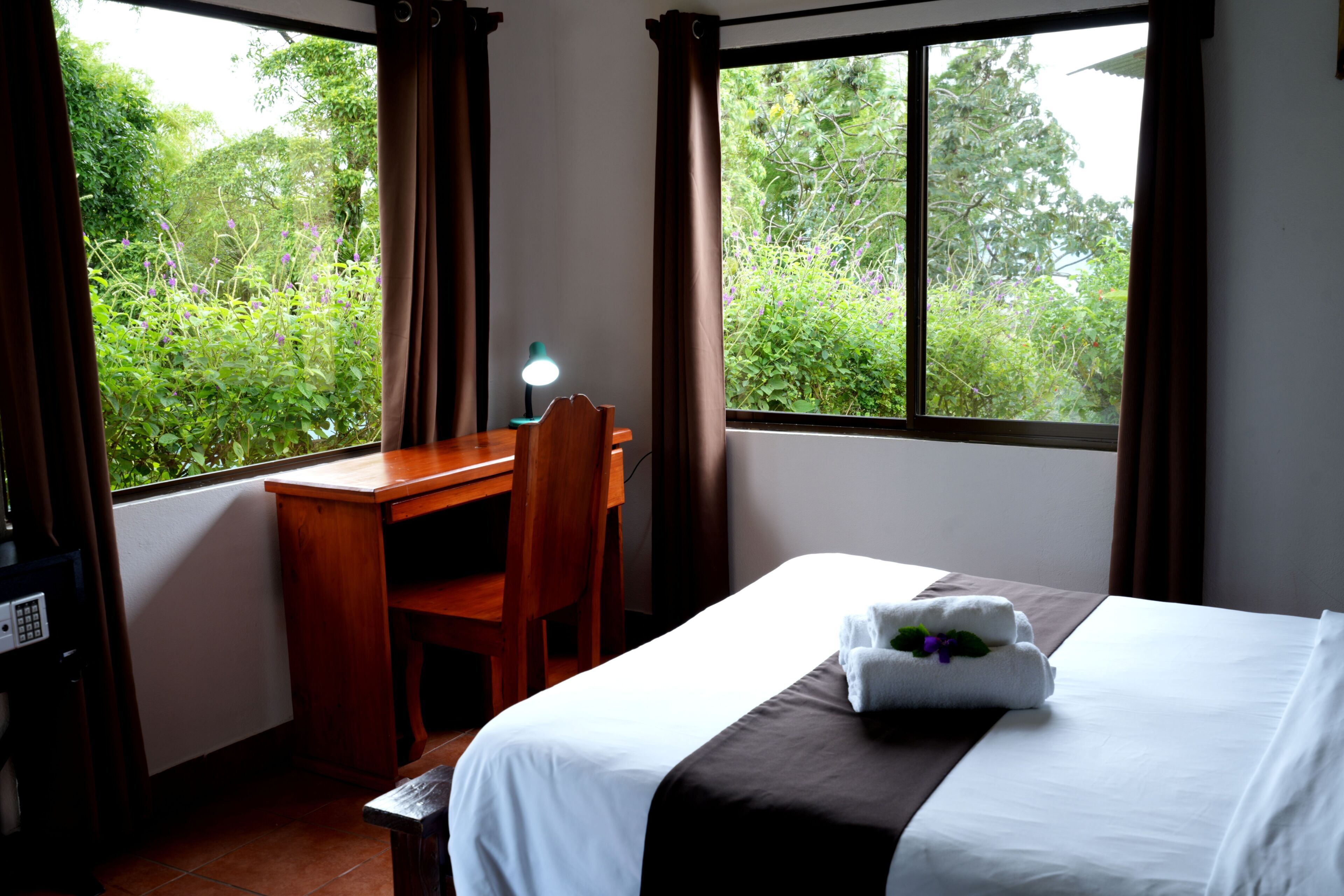 A hotel room with a bed, a desk, a chair, and a window with a view of greenery.