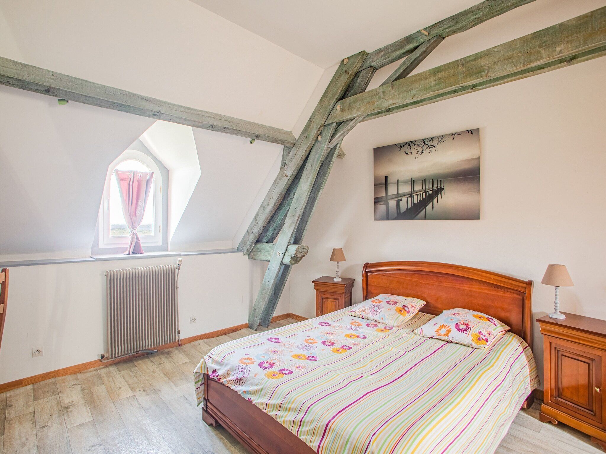 A bedroom with a sloped ceiling, a wooden bed, a radiator, a window with curtains, and a painting on the wall.