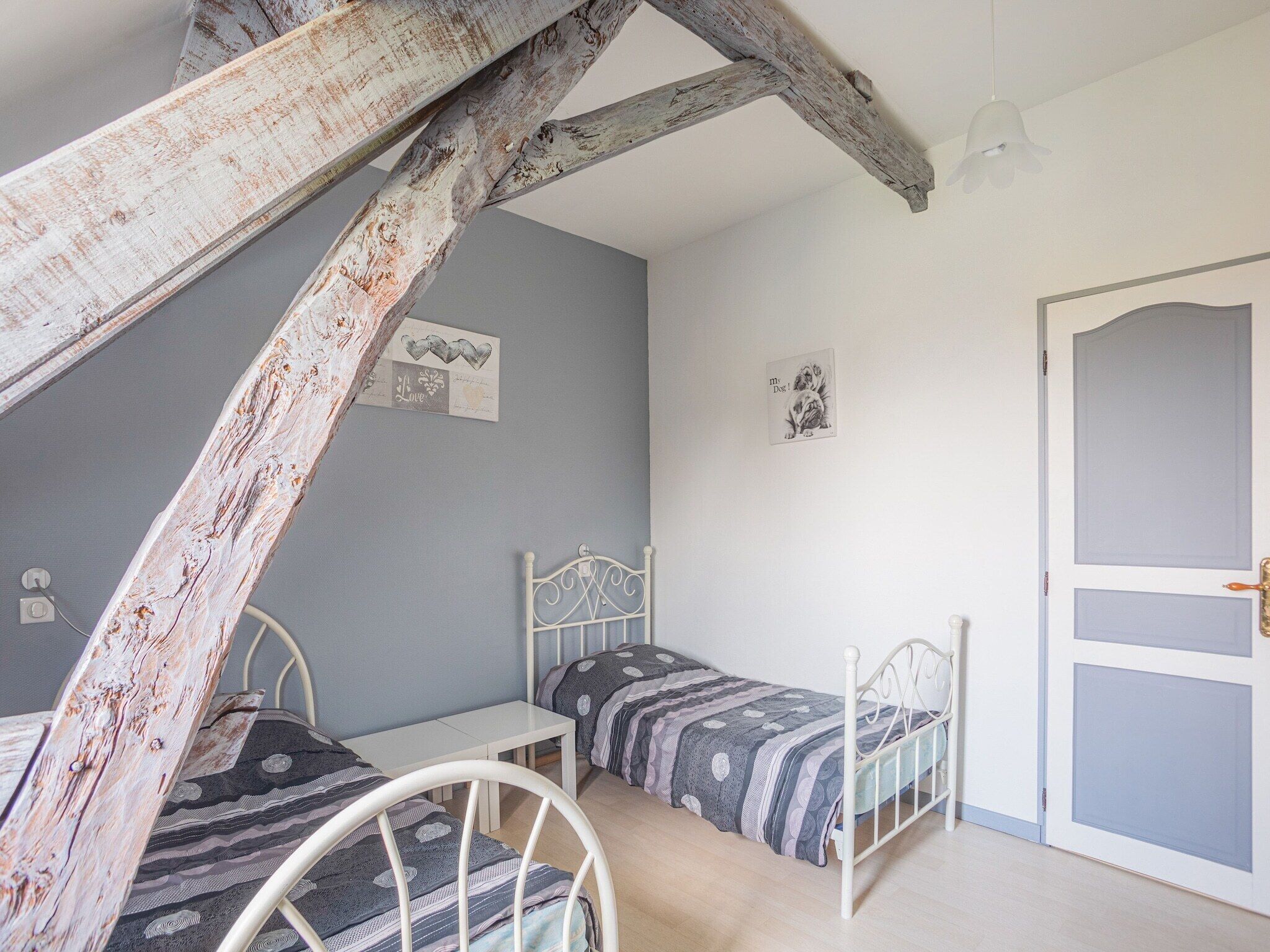 A bedroom with two single beds, a small table, a chair, a wooden beam ceiling, and a door.