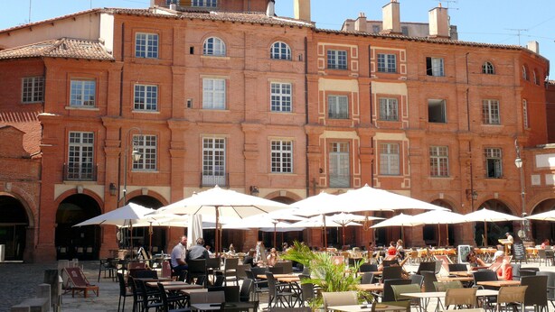 An outdoor cafe with tables and chairs under umbrellas, set against a red brick building with multiple windows and arches.