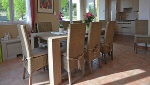 A dining area with wicker chairs and a wooden table, a kitchen in the background, and large windows with curtains.
