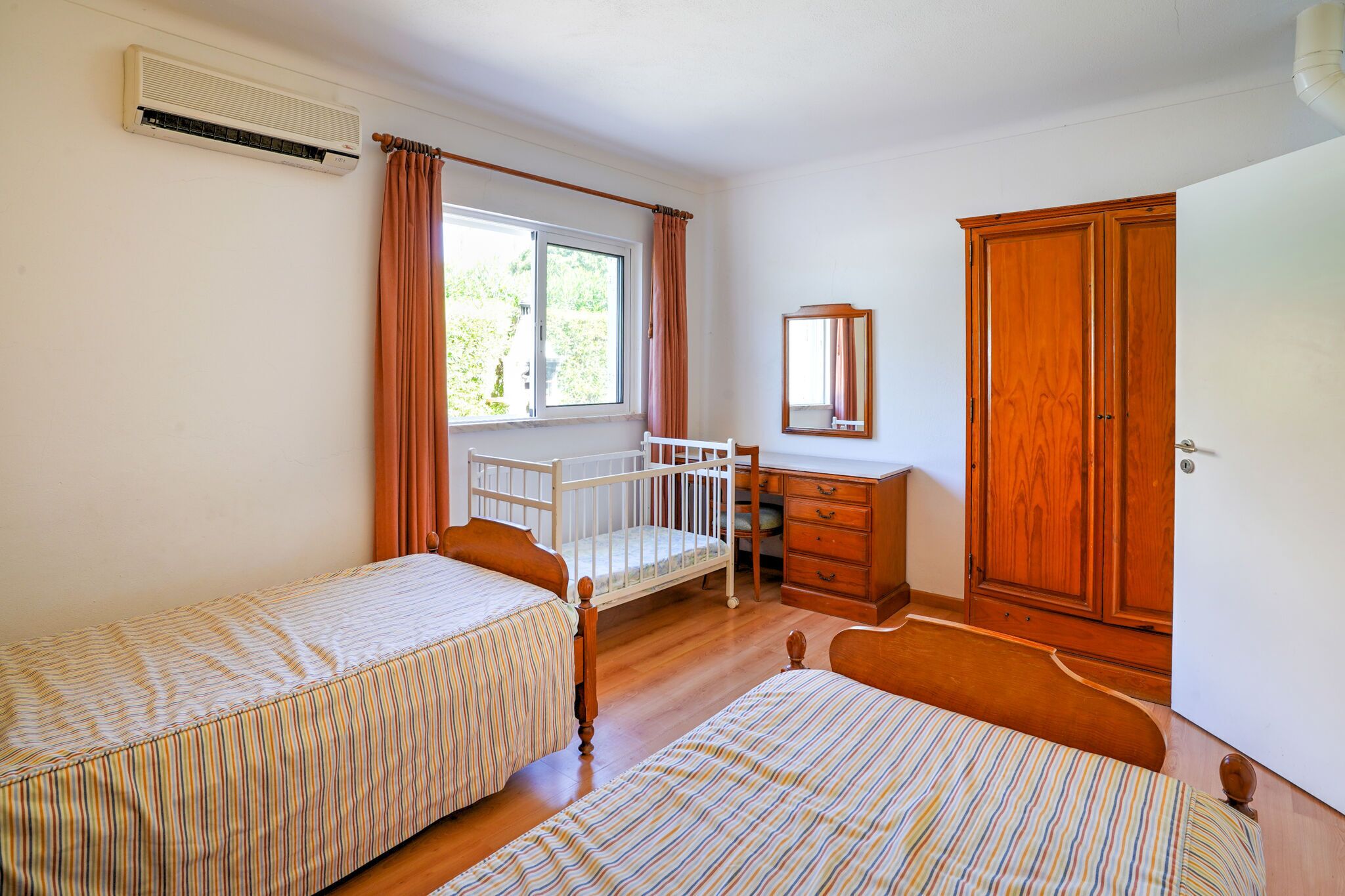 A bedroom with two single beds, a wooden wardrobe, a small dresser, a mirror, and an air conditioning unit.