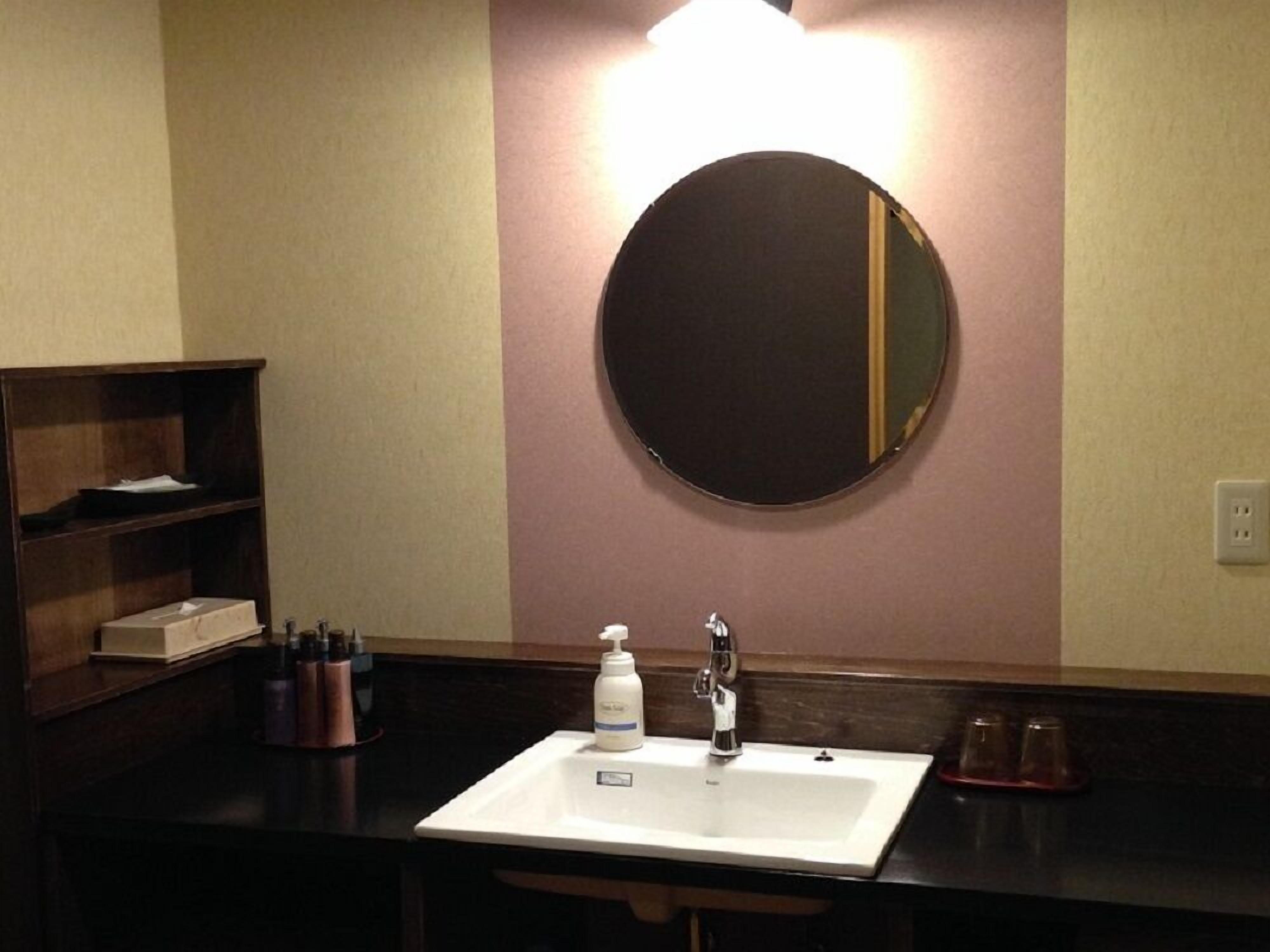 A hotel bathroom with a white sink, a round mirror, and a shelf with toiletries.