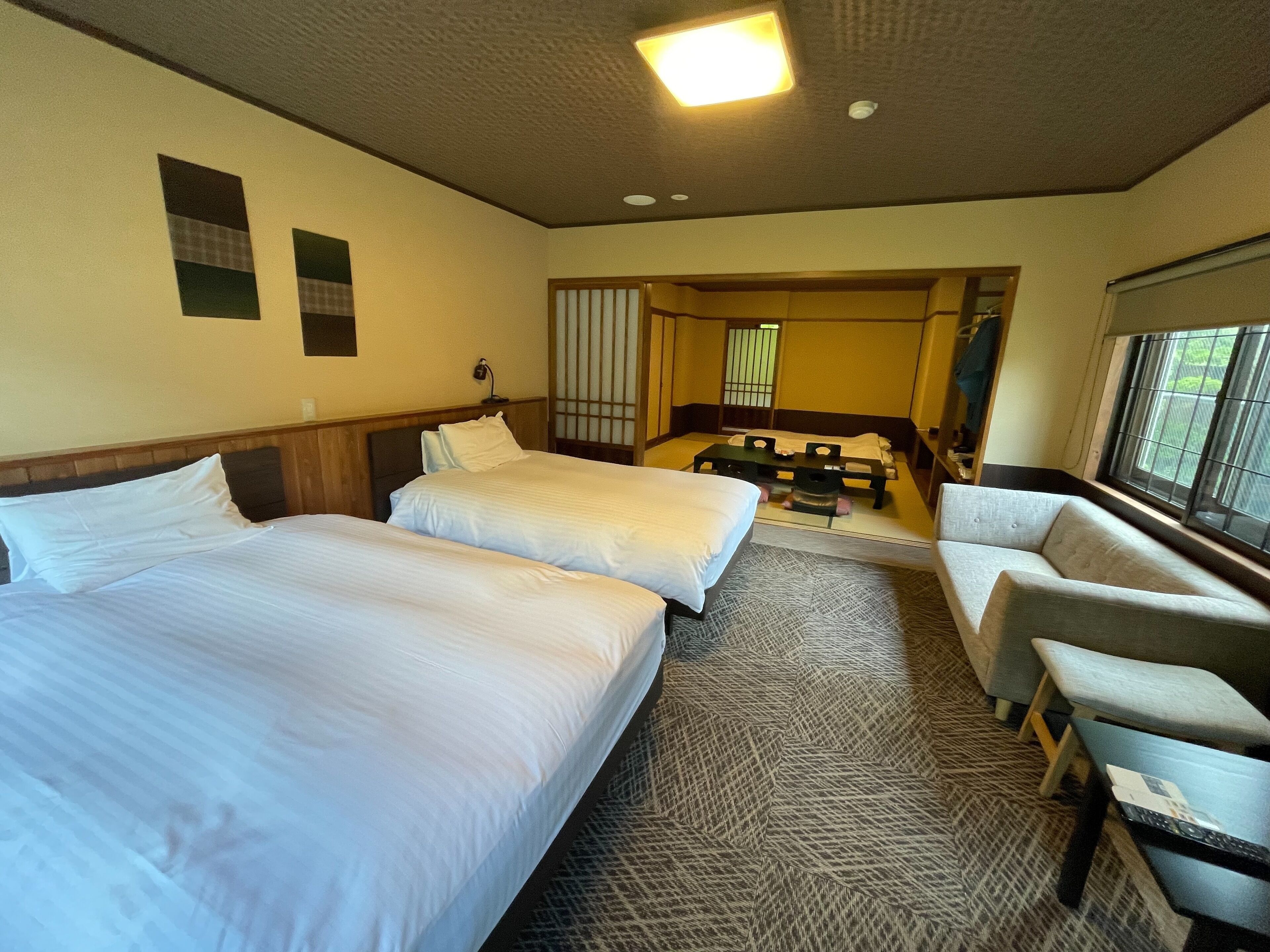 A hotel room with two beds, a sofa, a small table, and a view of the outside through a window.