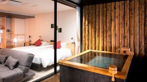 A modern hotel room with a large bed, a gray sofa, a wooden bathtub, and a wooden wall.