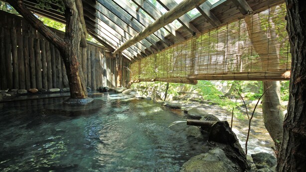 A traditional wooden bathhouse with a clear water pool, bamboo blinds, and a natural stone setting.