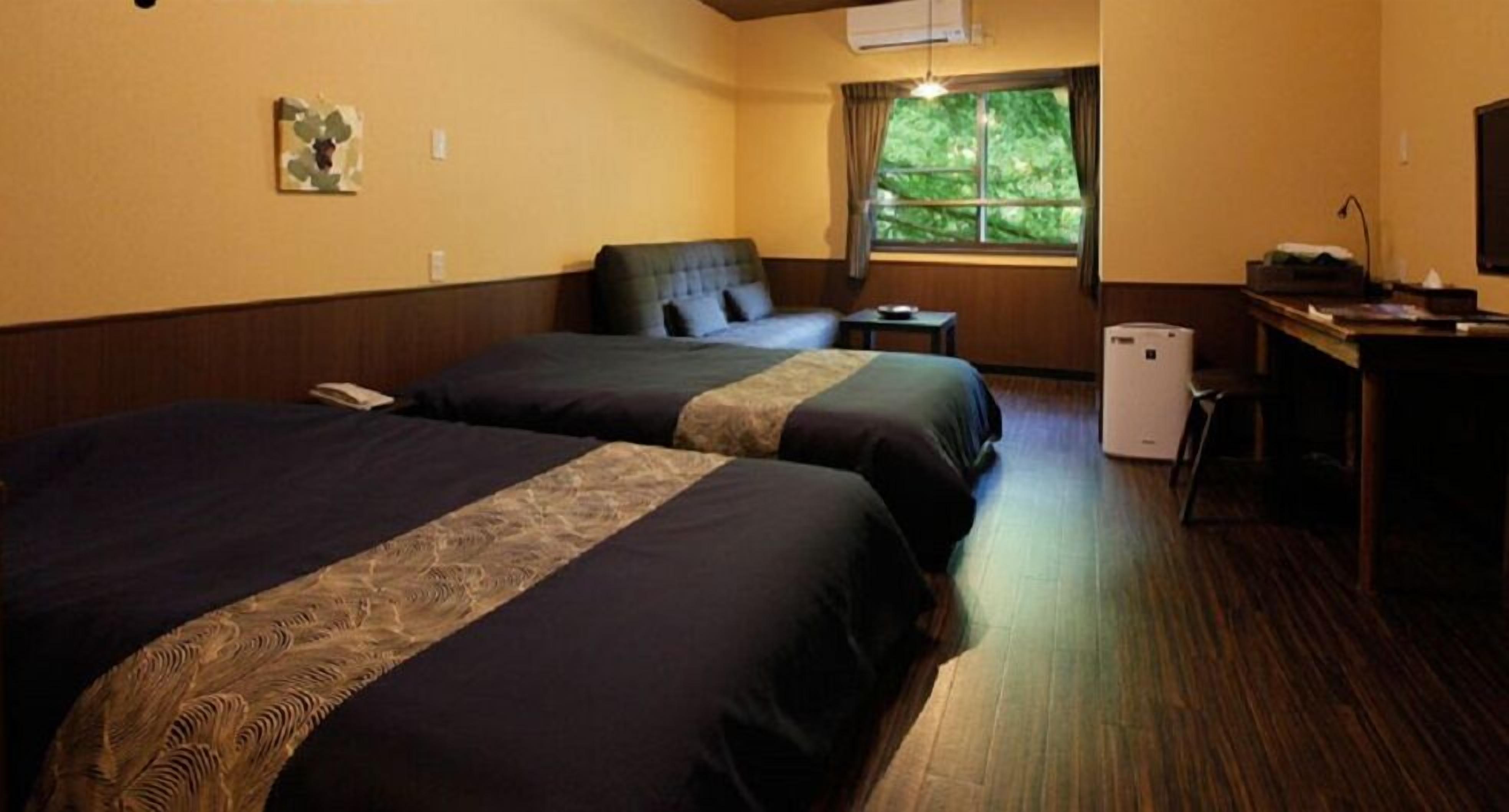 Habitación de hotel con dos camas, un escritorio, una silla, un ventanal con vista a árboles y un equipo de aire acondicionado.