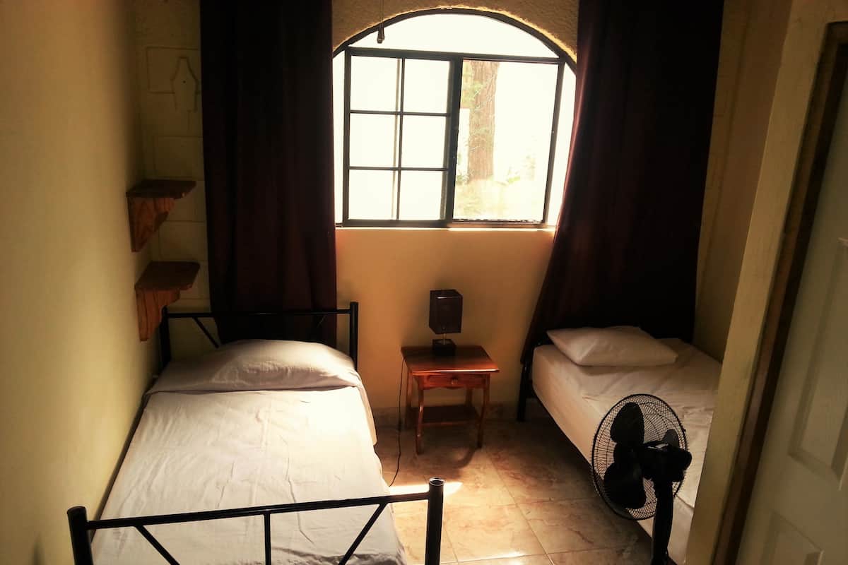 Una habitación pequeña, con poca luz, con dos camas individuales, una mesita con una lámpara y una ventana con cortinas.