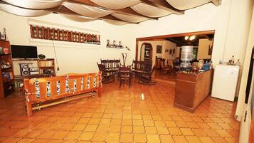 A spacious living area with a tiled floor, a wooden bench, a dining table with chairs, a television, and a bookshelf.