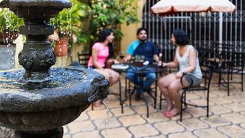 Una fuente de piedra con agua brotando de la parte superior, rodeada de mobiliario exterior con mesas y sillas, y plantas en macetas al fondo.