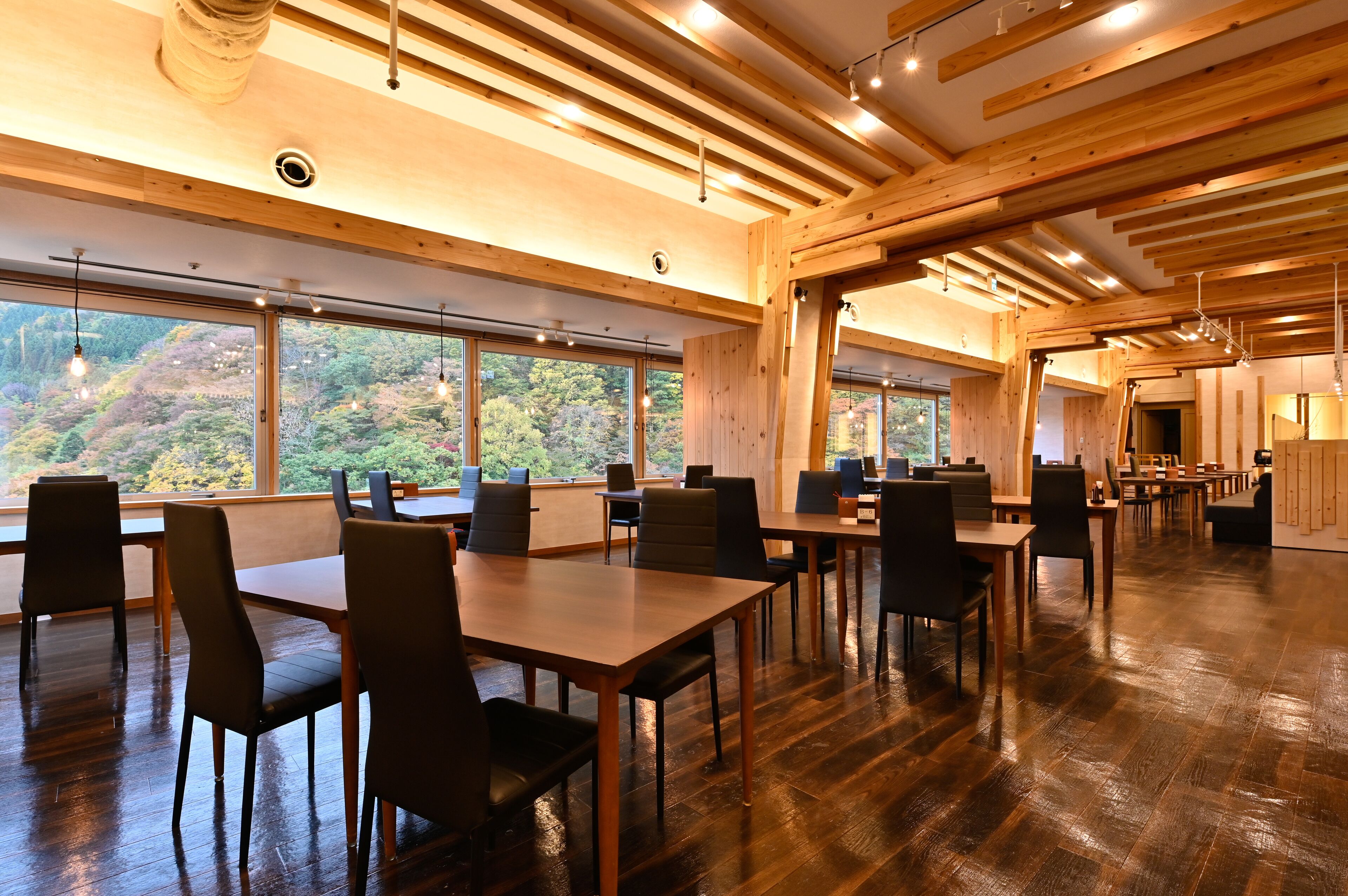 A spacious dining area with wooden tables and chairs, large windows offering a view of a forested hillside.