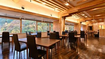 A spacious dining area with wooden tables and chairs, large windows offering a view of a forested hillside.