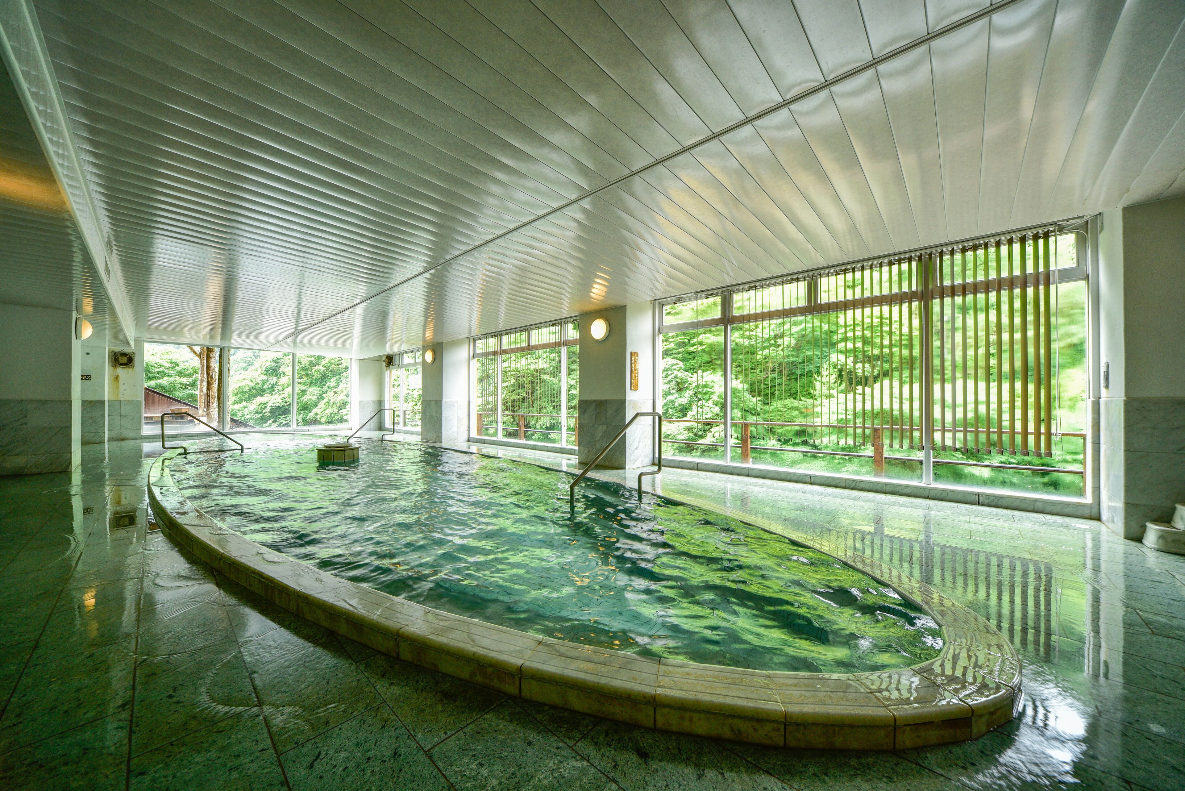 Una piscina interior con suelo de baldosas, ventanas amplias y techo con vigas horizontales.