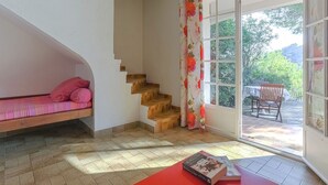 A room with a wooden staircase, a red table with a book, a pink sofa, and a window with a view of greenery.
