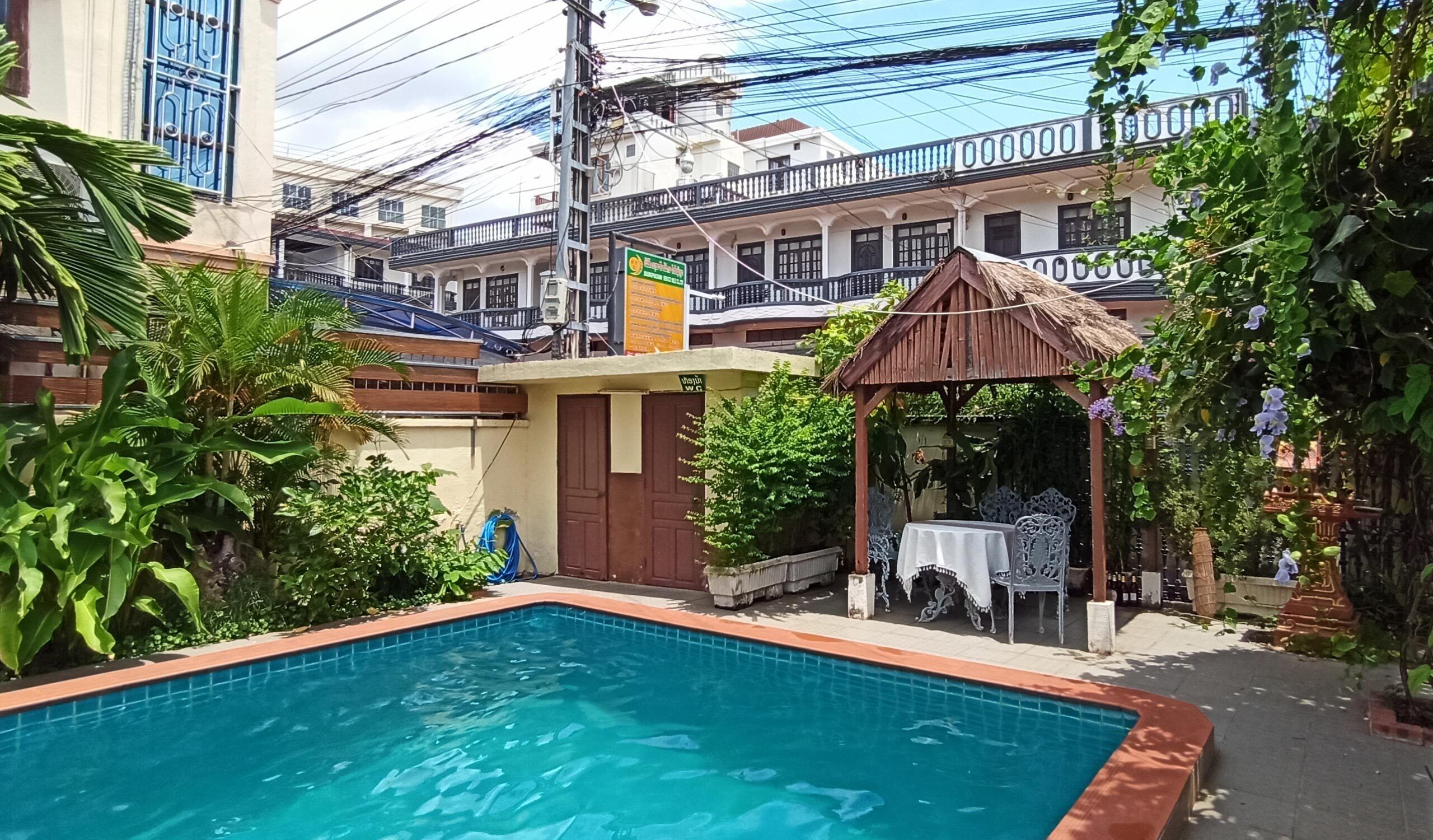 Piscina con un pabellón y asientos al aire libre, rodeada de abundante vegetación y edificios residenciales.