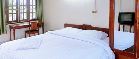 Een net opgemaakt bed met witte lakens, een houten hoofdbord, een klein houten bureau met een stoel, een televisie op een houten dressoir en een raam met groene gordijnen.