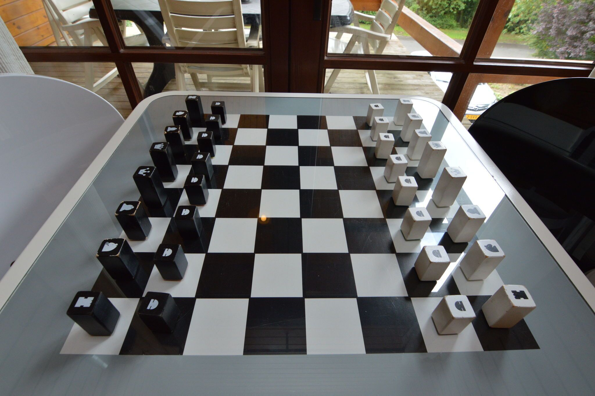 A checkered game board with black and white pieces, set on a table in a room with large windows.