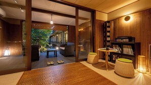 Un interior moderno con suelo de madera, un vidrio que da a un espacio exterior, una mesita con lámpara y una estantería.