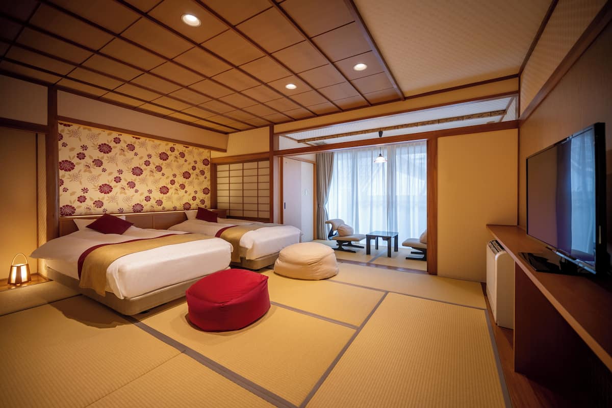 A hotel room with a traditional Japanese style, featuring tatami flooring, a large bed with multiple pillows, a red bean bag, a television, and a sliding door leading to a balcony.