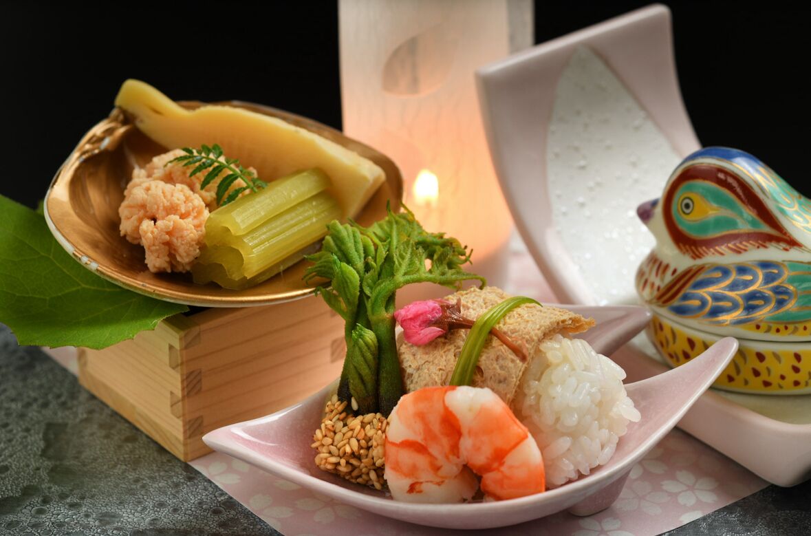 Refeição tradicional japonesa com arroz, camarão e legumes, servida em bandeja de madeira com uma pequena figura decorativa de pássaro.