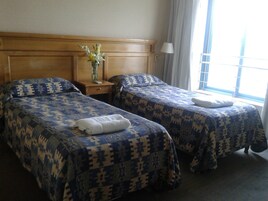 Quarto de hotel com duas camas, cabeceira de madeira, mesa de cabeceira com um vaso e janela com cortinas.