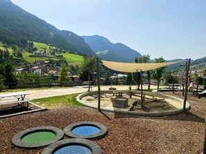 Une aire de jeux dotée d’une structure d’escalade circulaire, de tables de pique-nique et offrant une vue sur un village en arrière-plan.