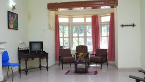 Una sala de estar con un televisor sobre una mesa de madera, dos sillones estampados, una silla azul y un ventanal con cortinas rojas.