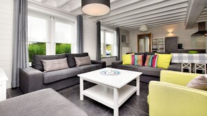 A modern living room with a white coffee table, grey sofas, and a yellow armchair. There is a kitchen area with a dining table and a large window. - Holiday home suitable for families with sauna (Wilogne)