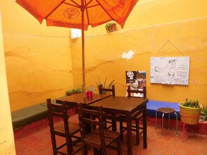 A small, enclosed dining area with a wooden table and chairs, an orange umbrella, a whiteboard with handwritten notes, and a small potted plant. - Cirque Hostel 2 (Lima)