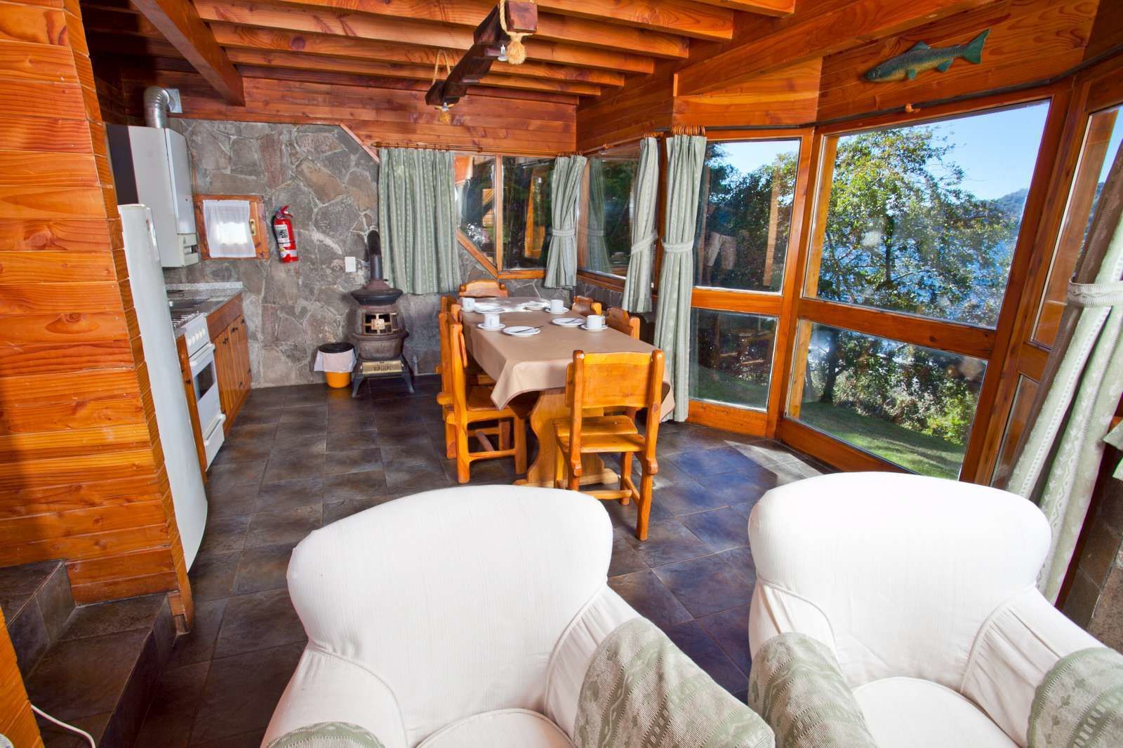 A cozy cabin interior with a stone wall, wooden ceiling, a dining table with chairs, a kitchen area, and large windows offering a view of the outdoors.