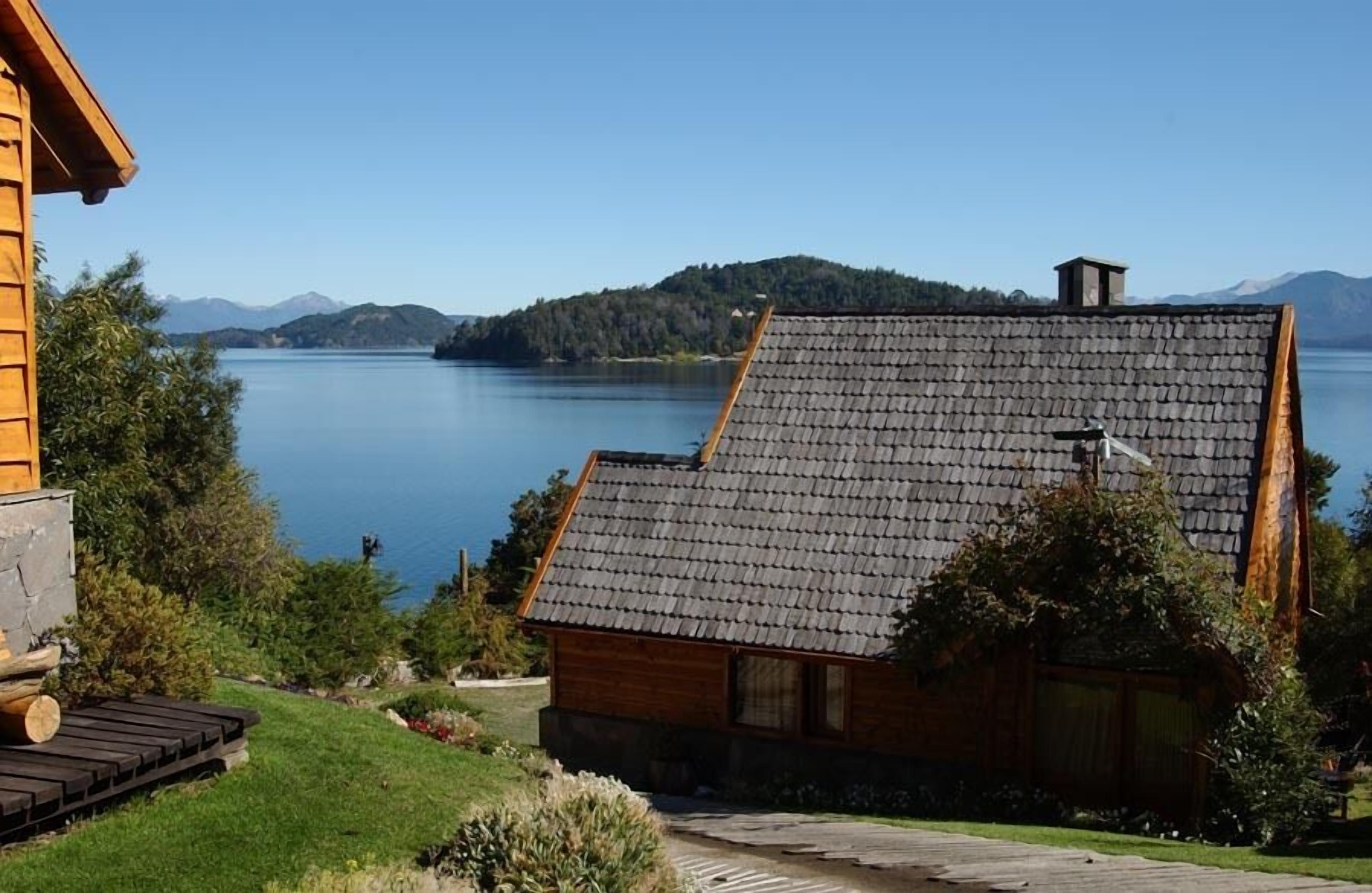 A wooden cabin with a sloped roof overlooking a lake, surrounded by greenery and mountains in the background.