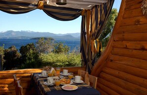 Una cabaña de madera con una mesa dispuesta para el desayuno, con vistas a un paisaje montañoso y un cuerpo de agua.