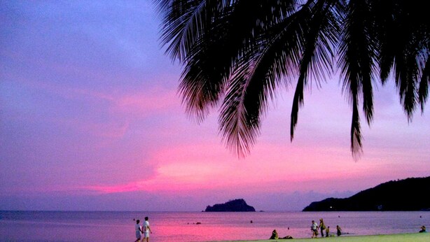A beach scene with people walking and sitting on the sand, palm trees, and a sunset over the water. - Apartamento Mar Azul - Smr143a (Santa Marta)