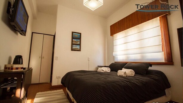 A bedroom with a bed, a television, a window with blinds, a painting, and a wooden ceiling. - Tower Rock Departamentos Estándar (Puerto Deseado)