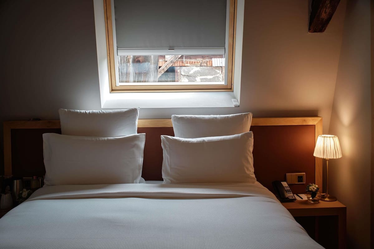 A hotel room with a bed, pillows, a nightstand, a lamp, and a window with blinds.