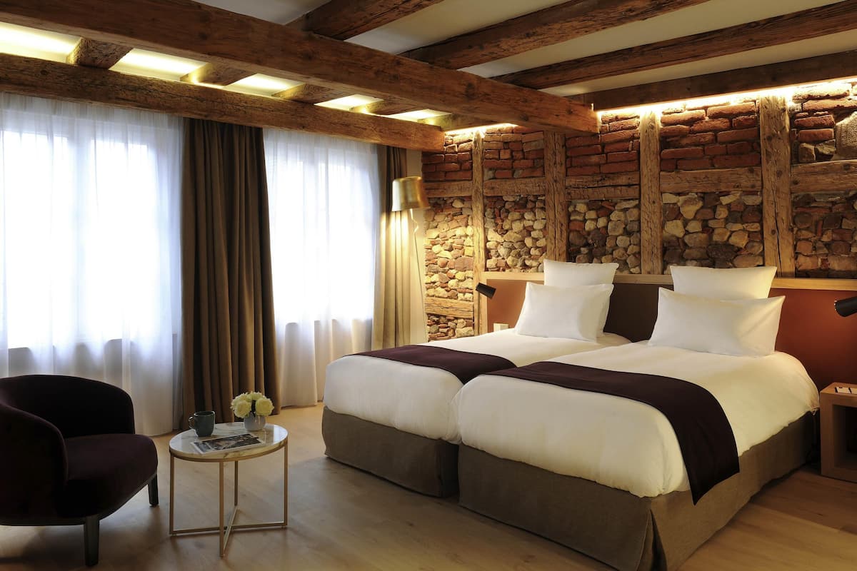 A room with a bed, a chair, a small table with flowers, and a stone wall.