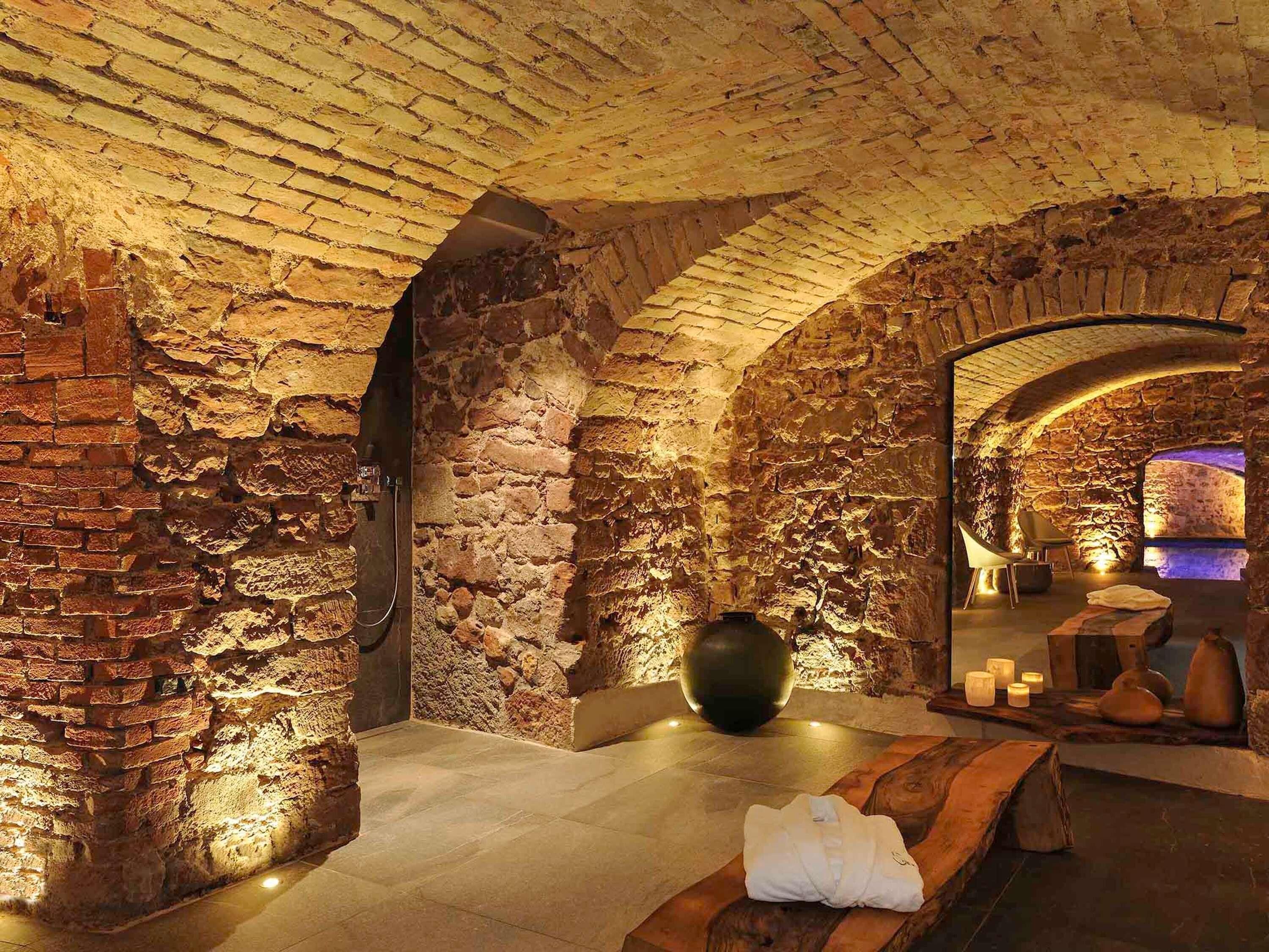 Una habitación con paredes de piedra, un jarrón grande, un banco de madera y una pequeña mesa con velas.