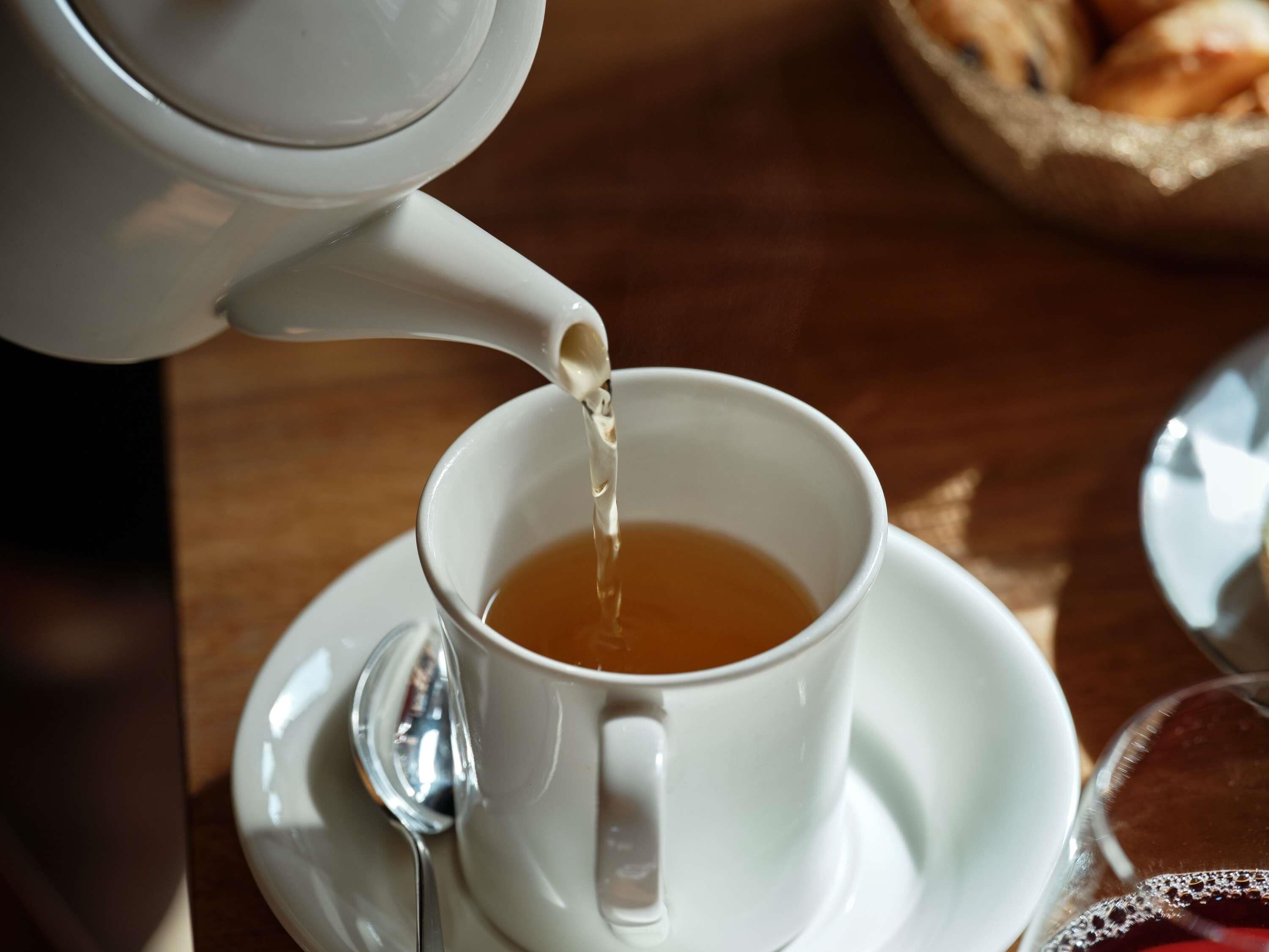 A white teapot pouring tea into a white cup on a saucer, with a spoon beside it.