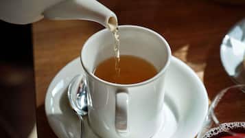 A white teapot pouring tea into a white cup on a saucer, with a spoon beside it.