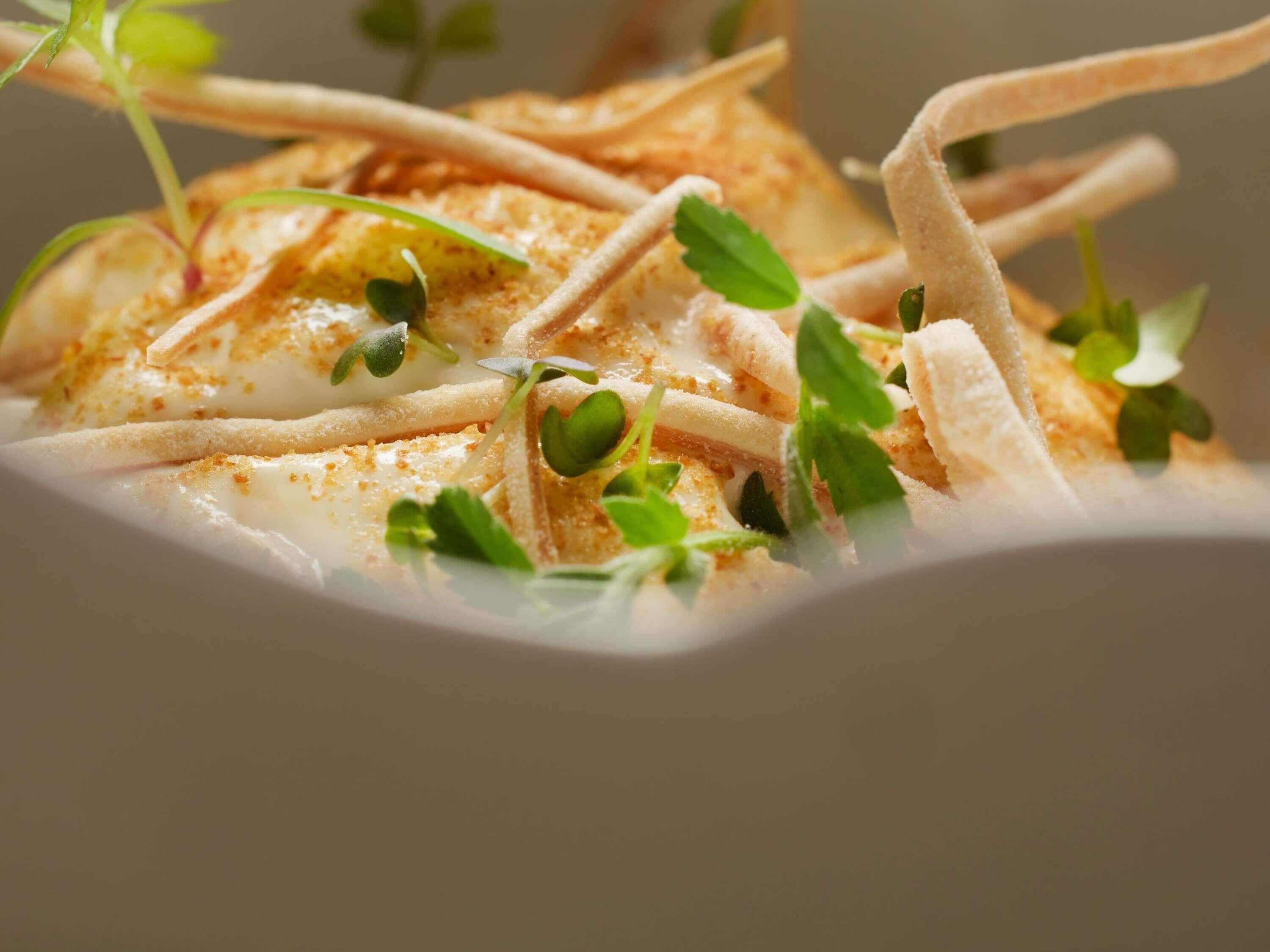 A close-up of a dish with thinly sliced ingredients, possibly fish, garnished with microgreens and a sprinkle of seasoning.